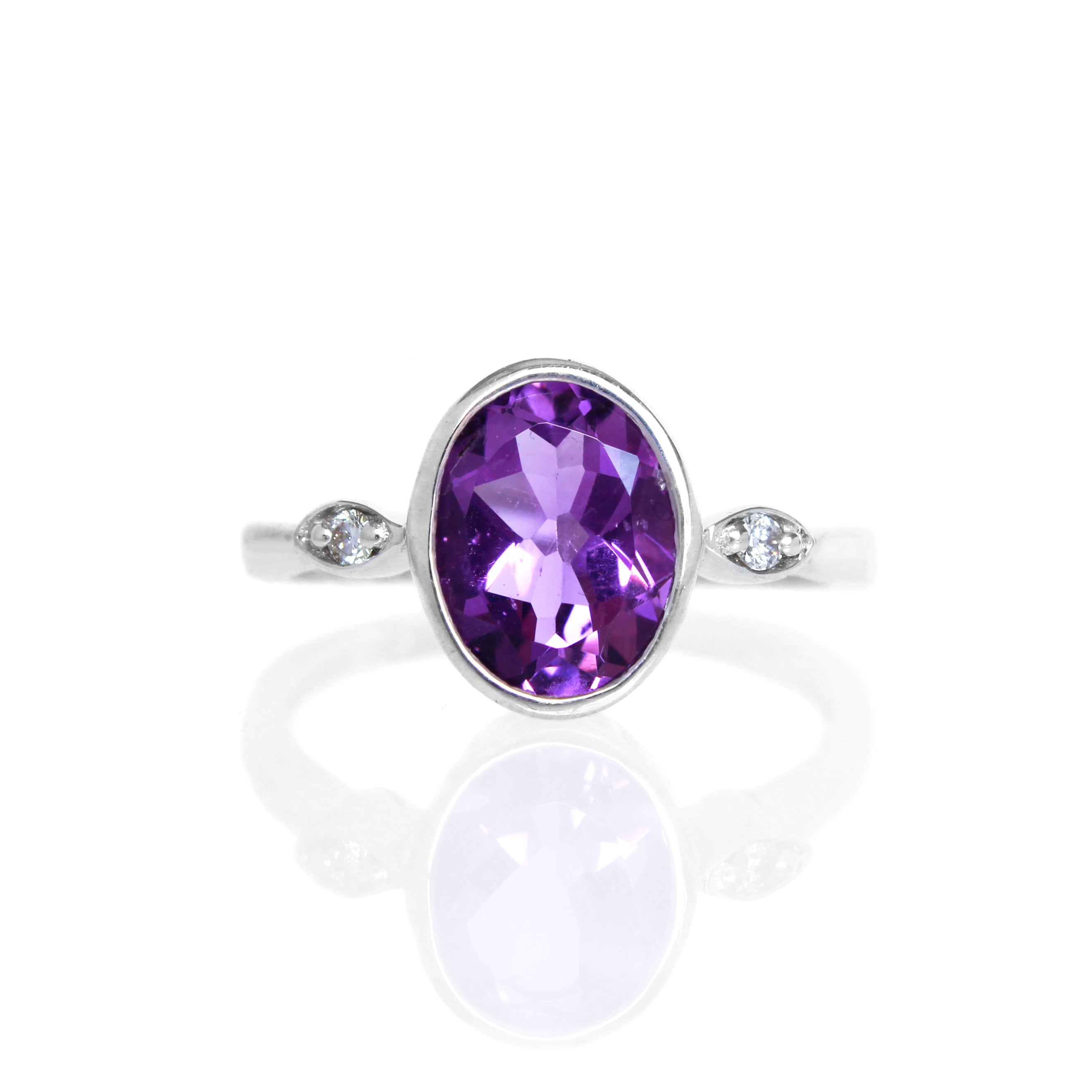 A product photo of a silver ring with a large bezel-set amethyst centre stone sitting on a white background. The silver band is simple and smooth, connecting on either side of a vertically-oriented oval-cut amethyst stone surrounded by a solid frame of silver. Before the band reaches the centre stone, it splits into two small horizontal silver oval frames on each side, each housing a small diamond.