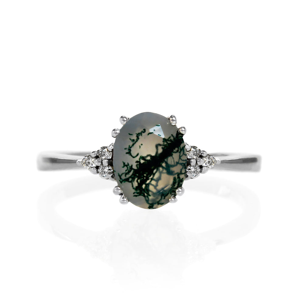 A product photo of a white gold moss agate and diamond trio ring sitting on a white background. The oval, naturally-included moss agate gemstone stands in stark contrast to the little clusters of three classic white diamond stones on either side.