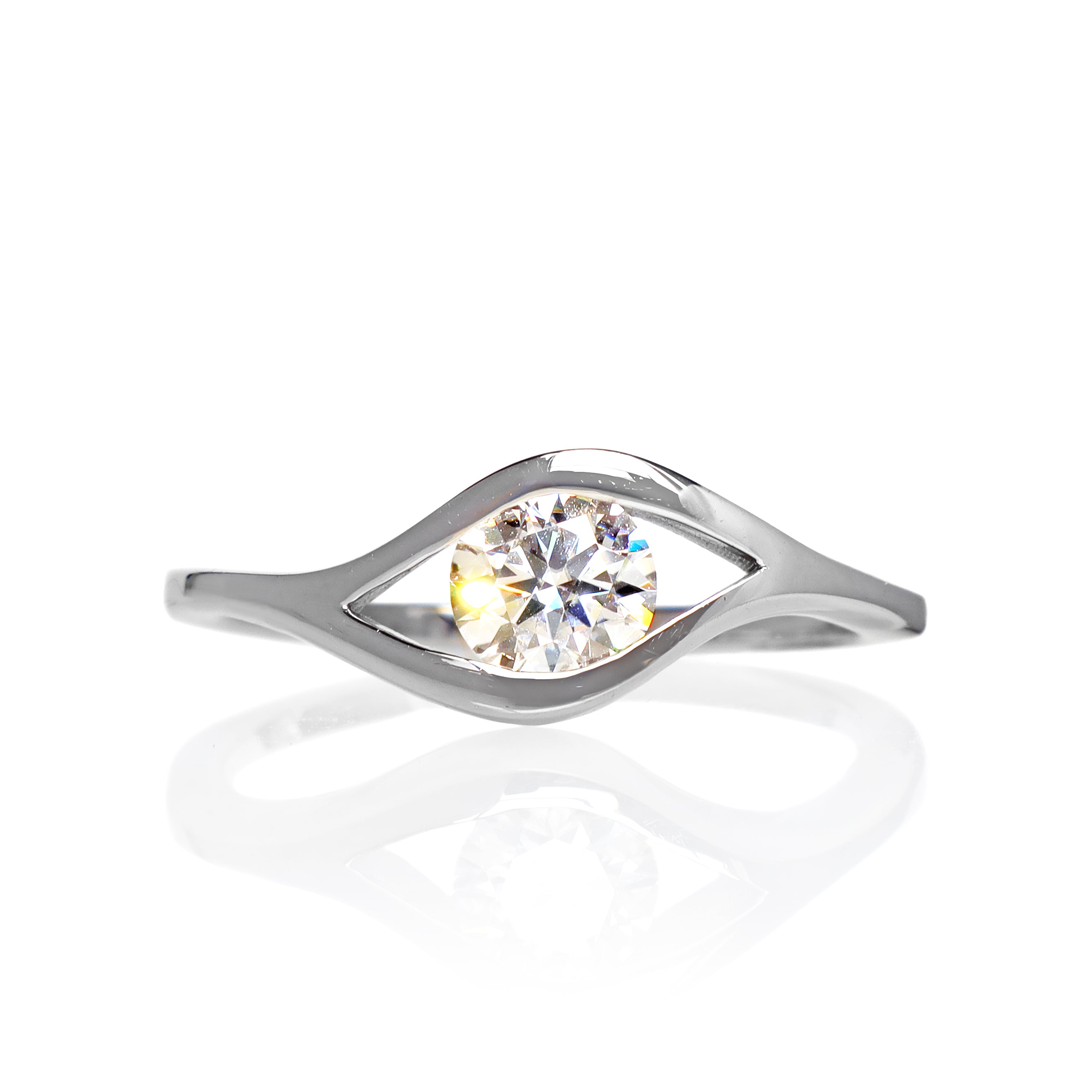 A product photo of moissanite ring in 9k white gold sitting on a clear white background. The reflective round-cut moissanite sits in the centre, and a simple white gold band splits on either side of the jewel, holding it in place like the corners of an eye.