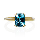 A product photo of a yellow gold london blue topaz ring sitting against a white background. The golden band is plain and smooth, contrasted by the large rectangular cushion-cut ocean blue stone in the centre.