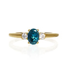A product photo of a yellow gold london blue topaz ring sitting against a white background. The golden band is plain and smooth, and the centre upright octagonal topaz stone is framed by a single white diamond on either side.