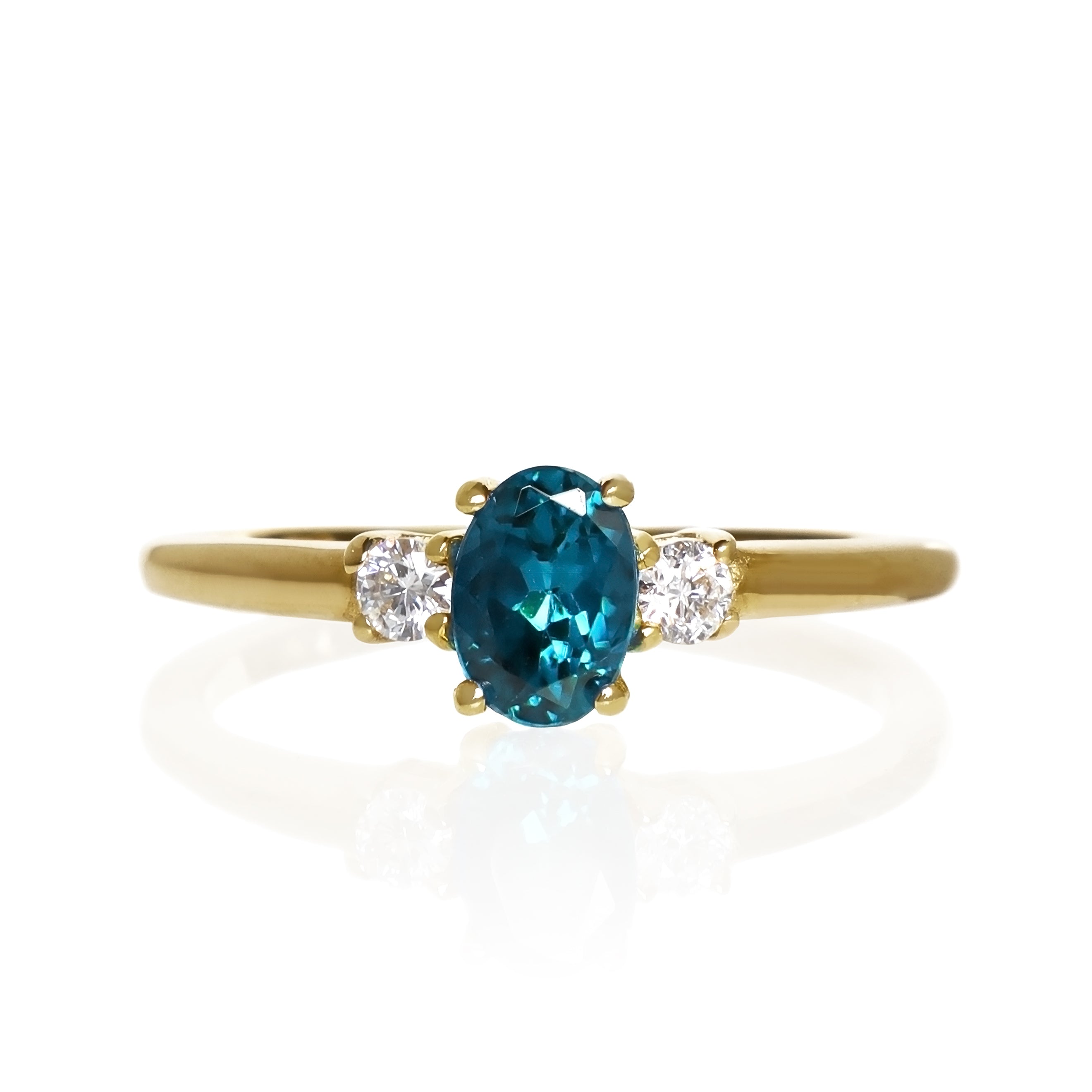 A product photo of a yellow gold london blue topaz ring sitting against a white background. The golden band is plain and smooth, and the centre upright octagonal topaz stone is framed by a single white diamond on either side.