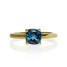 A product photo of a yellow gold london blue topaz ring sitting against a white background. The golden band is plain and smooth, contrasted by the large square cushion-cut ocean blue stone in the centre.