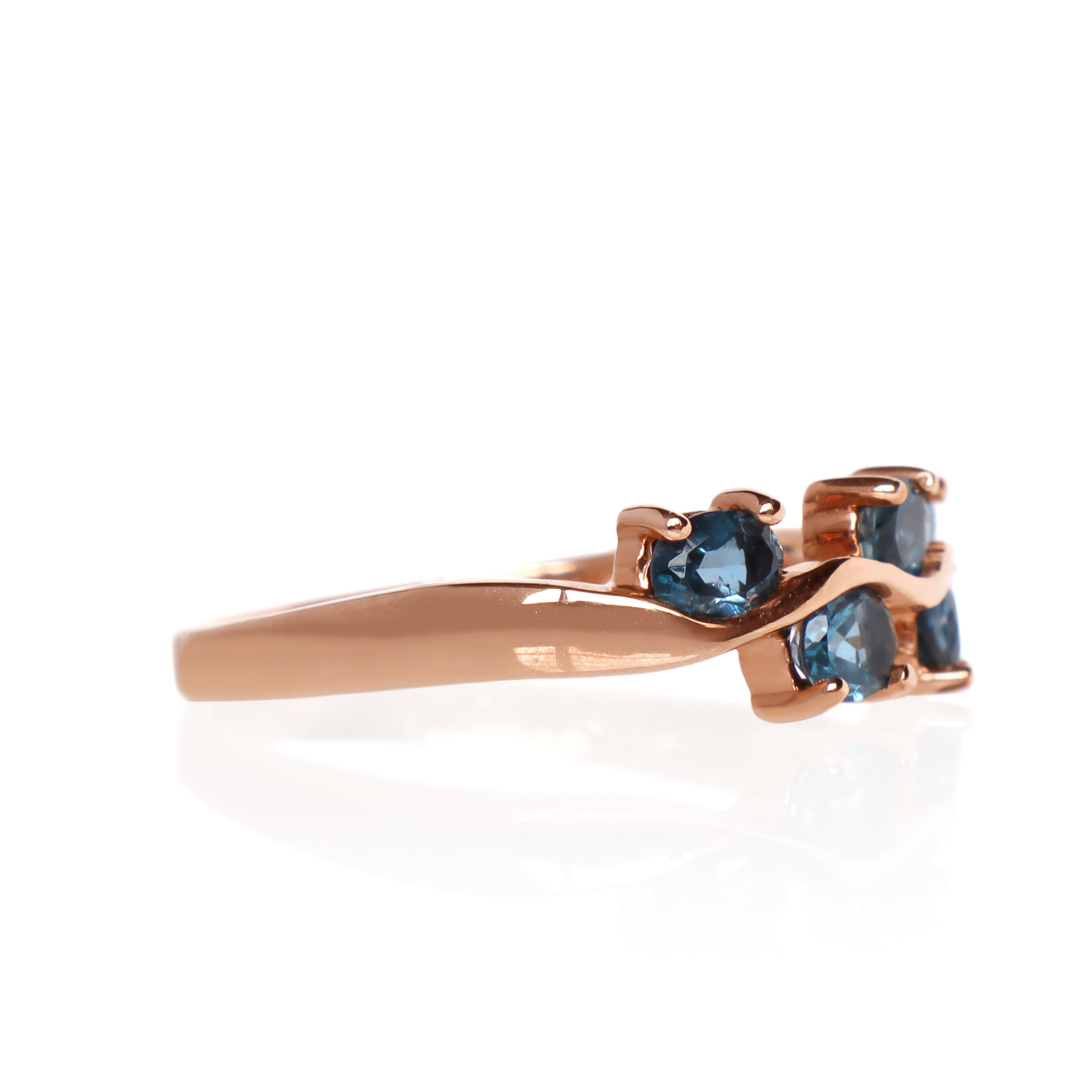 A product photo of a rose gold london blue topaz ring sitting against a white background. The rose gold band flows up and down like waves, with each rise and fall punctuated by a single oval-cut topaz stone, to a total of 5.
