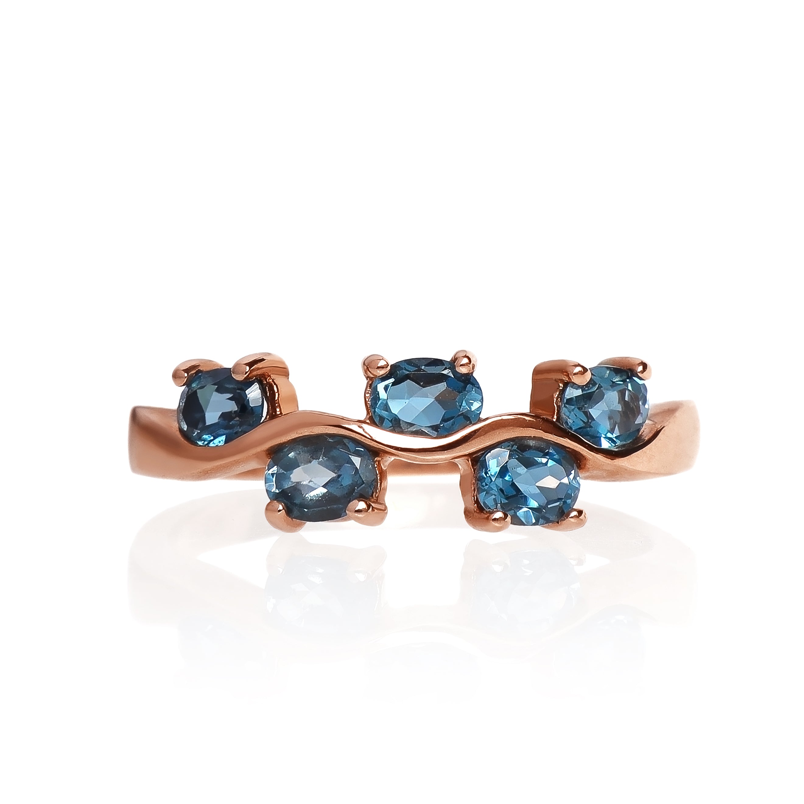 A product photo of a rose gold london blue topaz ring sitting against a white background. The rose gold band flows up and down like waves, with each rise and fall punctuated by a single oval-cut topaz stone, to a total of 5.