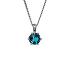 A product photo of a white gold london blue topaz pendant sitting against a white background. The highly-reflective circle cut topaz stone is held in place by 6 dainty white gold claws. It is suspended by a simple white gold chain.