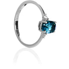 A product photo of a white gold london blue topaz ring sitting against a white background. The white gold band is plain and smooth, and the centre oval topaz stone is framed by a single white diamond on either side.