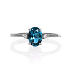 A product photo of a white gold london blue topaz ring sitting against a white background. The white gold band is plain and smooth, and the centre oval topaz stone is framed by a single white diamond on either side.