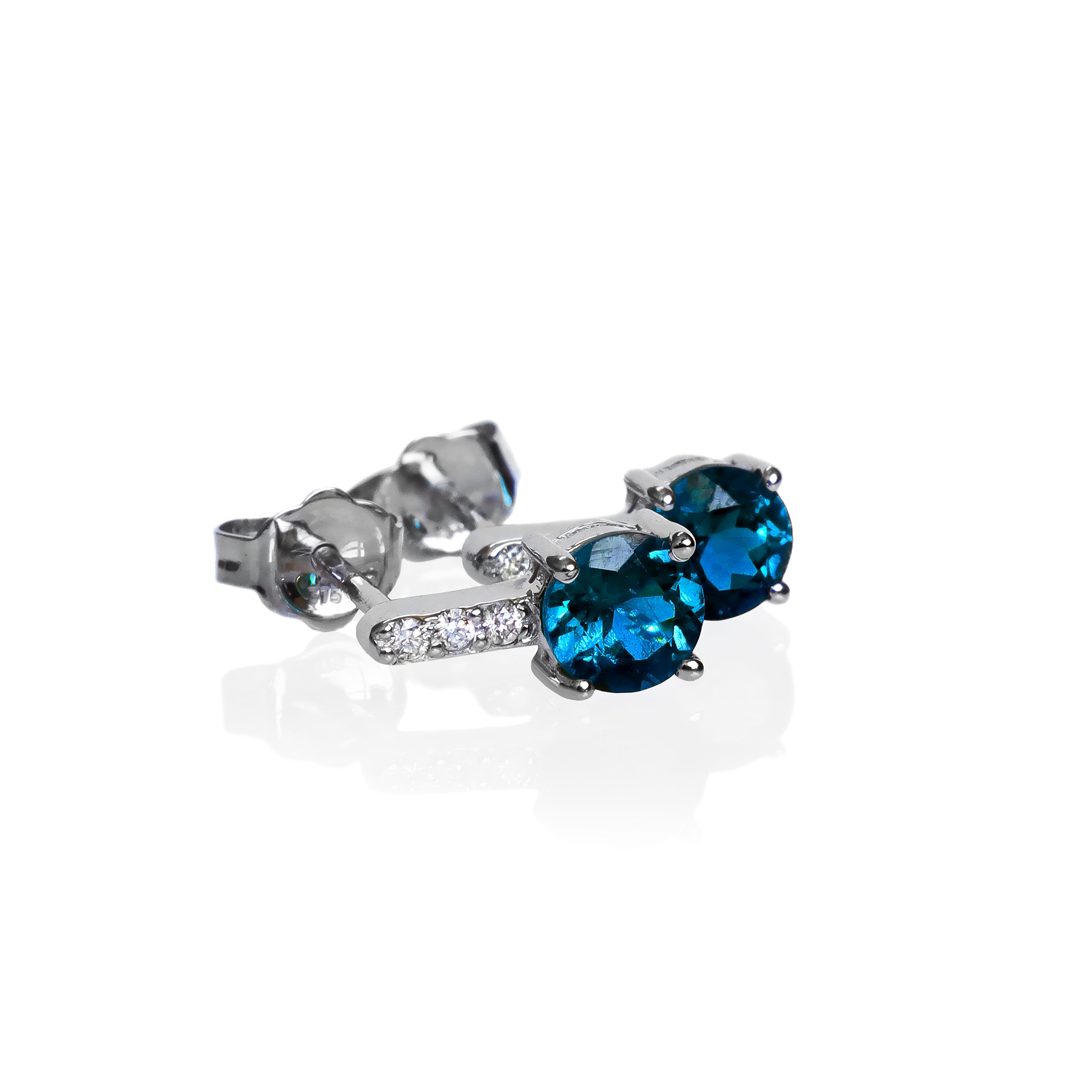 A product photo of a pair of white gold london blue topaz earrings sitting against a white background. The simple circle-cut stones are contrasted by the white diamond trio protruding in a straight line above each stone, encased in white gold.