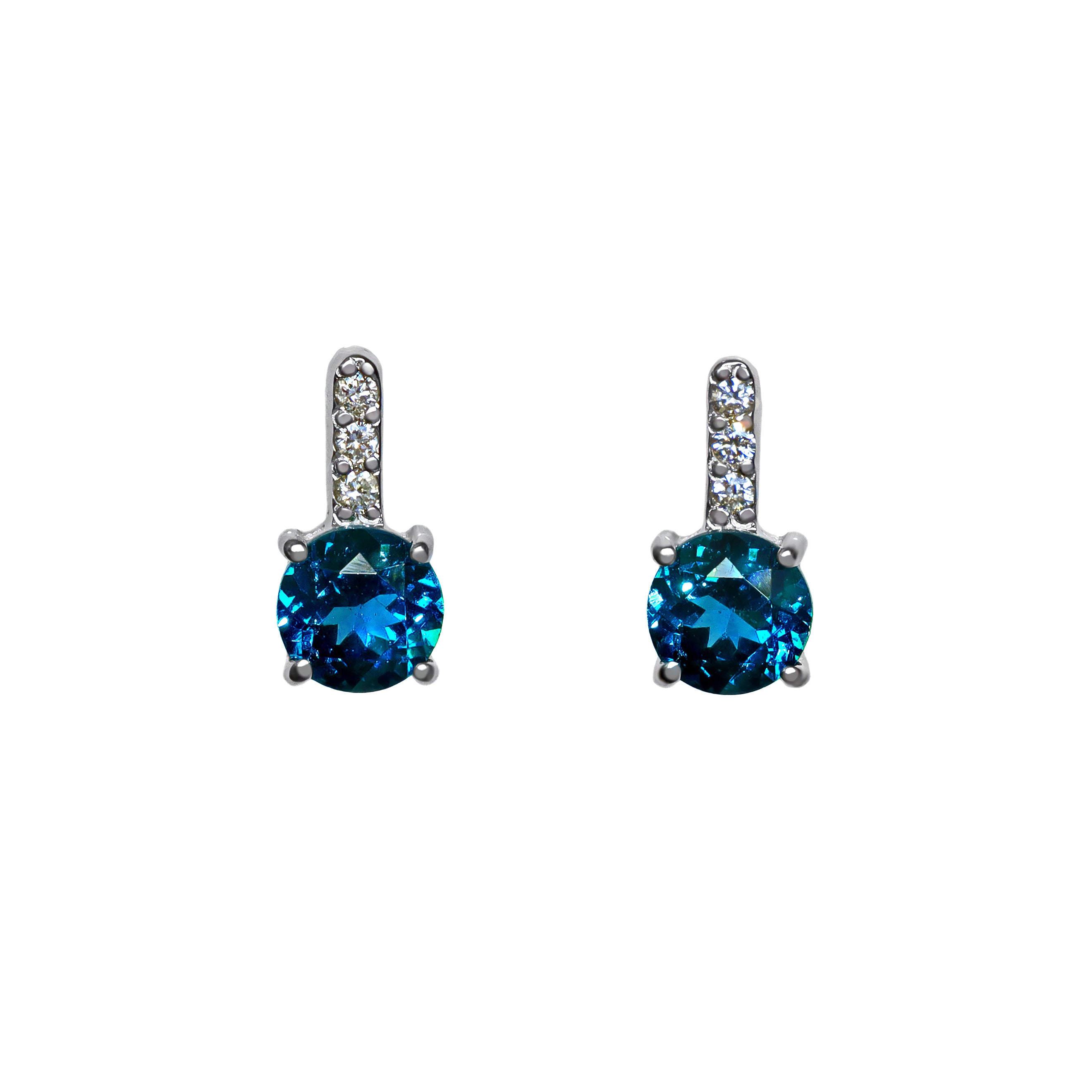 A product photo of a pair of white gold london blue topaz earrings sitting against a white background. The simple circle-cut stones are contrasted by the white diamond trio protruding in a straight line above each stone, encased in white gold.