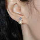 A product photo of delicate white pearl earring studs photographed on a model's earlobe. The white pearls each have aquamarine detailing above them connecting to the white gold studs.