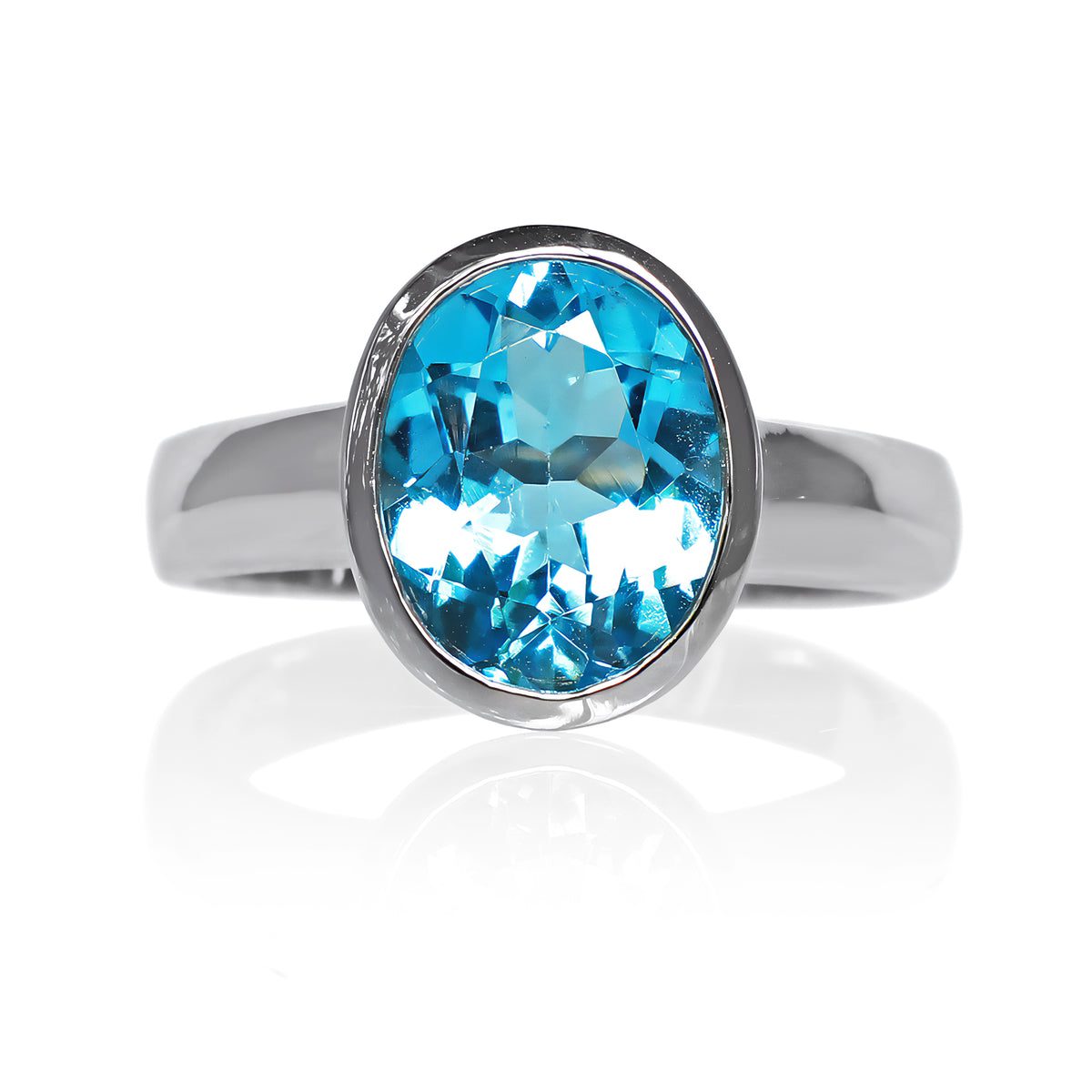 A product photo of a silver ring with a large bezel-set topaz centre stone sitting on a white background. The silver band is simple and smooth, connecting on either side of a vertically-oriented oval-cut topaz stone surrounded by a solid frame of silver. The topaz jewel is almost the colour of a tropical sea, reflecting bright blue light across its multi-faceted surface.