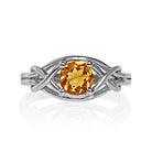 A product photo of an ornate silver ring with a circle-cut citrine centre stone sitting on a white background. The back of the silver band is simple and smooth, before splitting into four serpentine tendrils on either side that delicately coil around one another before meeting on either side of the circle-cut centre stone, which is held in place by four small silver claws. The citrine jewel is almost honey-coloured, reflecting a bright orangey yellow colour across its multi-faceted surface.