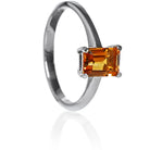A product photo of a silver ring with an emerald-cut citrine centre stone sitting on a white background. The silver band is simple and smooth, connecting on either side of a vertically-oriented emerald-cut citrine centre stone held in place by four silver claws. The citrine jewel is almost honey-coloured, reflecting a warm orangey yellow colour across its multi-faceted surface.