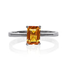 A product photo of a silver ring with an emerald-cut citrine centre stone sitting on a white background. The silver band is simple and smooth, connecting on either side of a vertically-oriented emerald-cut citrine centre stone held in placfe by four silver claws. The citrine jewel is almost honey-coloured, reflecting a warm orangey yellow colour across its multi-faceted surface.