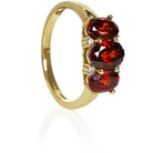A product photo of a Garnet Trio & Diamond Ring in Yellow Gold sitting on a plain white background. The largest garnet centrestone measures 7x5mm, with 4 tiny diamonds nestled against it, two on each side. The garnet stones are deep red, reflecting sanguine hues across their multi-faceted edges.