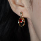 A product photo of 5.5mm Round Bezel-set Garnet Earring Studs in 9k Yellow Gold photographed on a model's earlobe. The bezel-set stones are nestled at the top of golden oval loops of similar thickness to the bezel frames. The garnets reflect sanguine hues across their multi-faceted edges.
