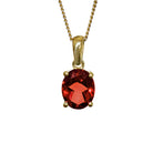 A product photo of a 9x7mm Oval Garnet Pendant in 9k Yellow Gold suspended against a white background. The impressively large and deeply-coloured oval-cut stone is contrasted by its overall minimalistic design, 4 simple golden claws holding the gem in place. It is suspended by a simple gold chain. The stone is a deep red, reflecting sanguine hues across its multi-faceted edges.