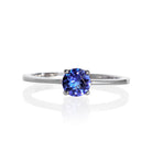 A product photo of a white gold tanzanite solitaire ring sitting on a white background. The band is smooth and simple, with the centre stone held in place by 4 small claws. The deep blue tanzanite stone reflects violet blue and indigo colours from its many edges.