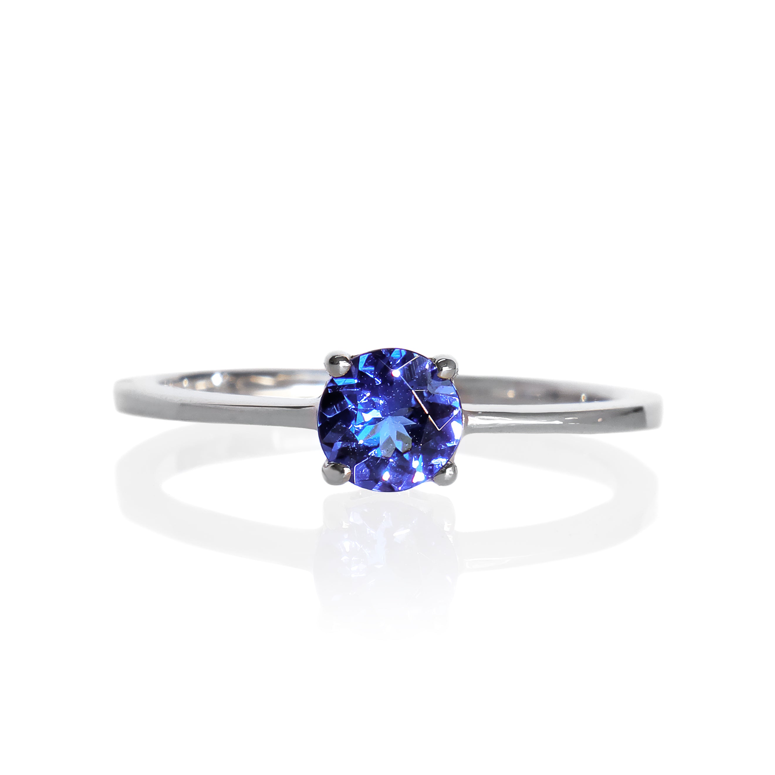 A product photo of a white gold tanzanite solitaire ring sitting on a white background. The band is smooth and simple, with the centre stone held in place by 4 small claws. The deep blue tanzanite stone reflects violet blue and indigo colours from its many edges.