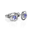 A product photo of white gold stud tanzanite earrings sitting on a white background. The small stones are framed by ornate, flowery white gold backs. The deep blue tanzanite stones reflect violet blue and indigo light from their many edges.