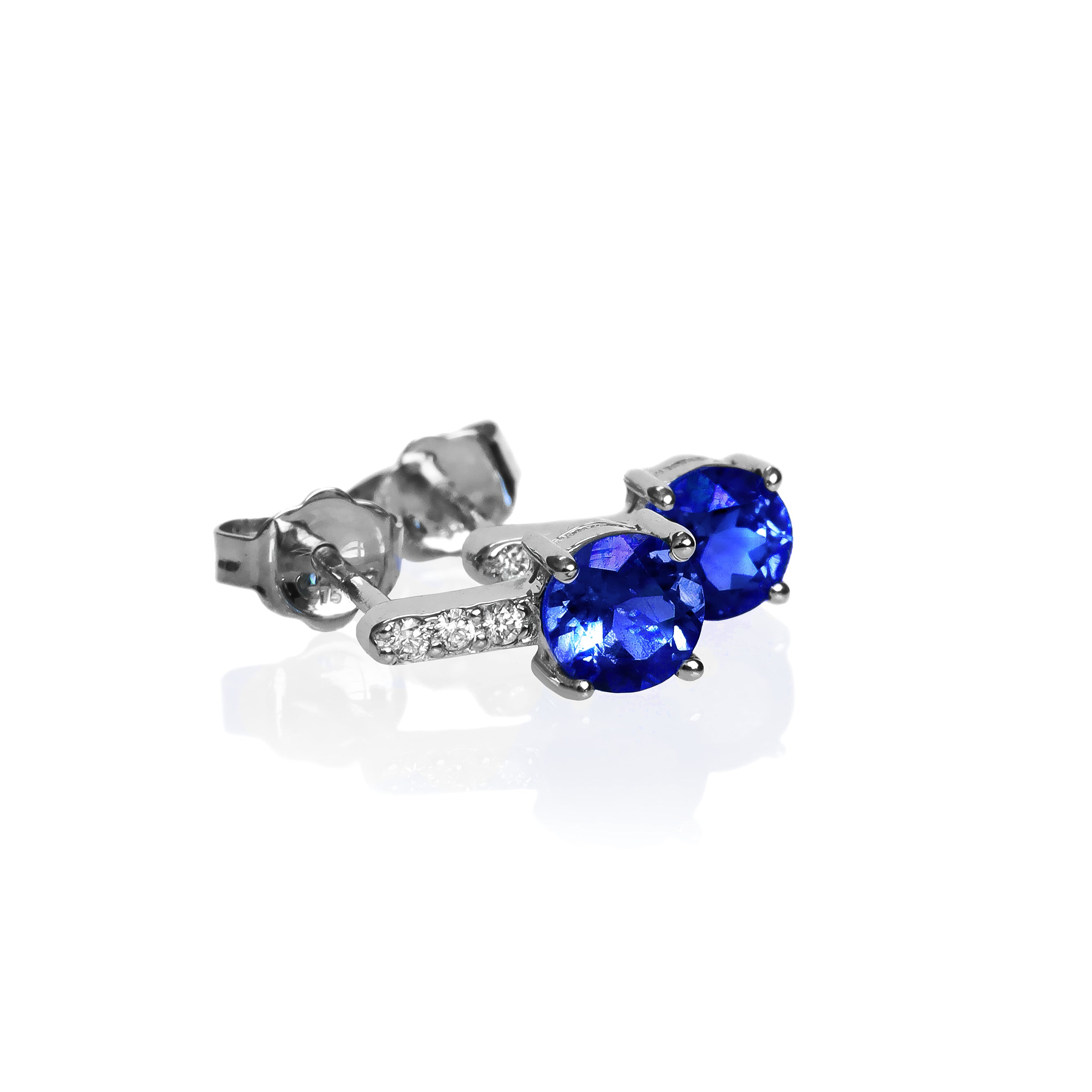 A product photo of white gold tanzanite stud earrings sitting on a white background. A golden strip connects the round stones to the studs, each strip adorned with 3 diamonds each. The deep blue tanzanite stones reflect violet blue and indigo light from their many edges.