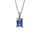A product photo of a white gold tanzanite pendant suspended by a chain against a white background. The emerald-cut tanzanite stone is held in place by 4 golden claws, and reflects violet blue and indigo colours from its edges.