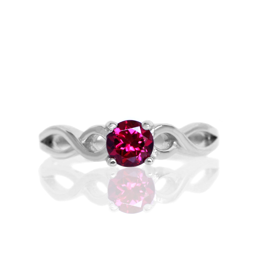 A product photo of an ornate silver ring with a rhodalite centre stone sitting on a white background. The silver band splits halfway along its length, becoming twisting and serpentine in appearance before meeting on either side of the dazzling plum-coloured 5mm stone, which is held in place by 4 silver claws.