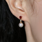 A product photo of a pair of ornate pearl drop stud earrings in yellow gold photographed on a model's earlobe. The pearls are creamy white and round, with one small diamond followed by a pear-shaped ruby on each piece linking them to the back of the golden stud.