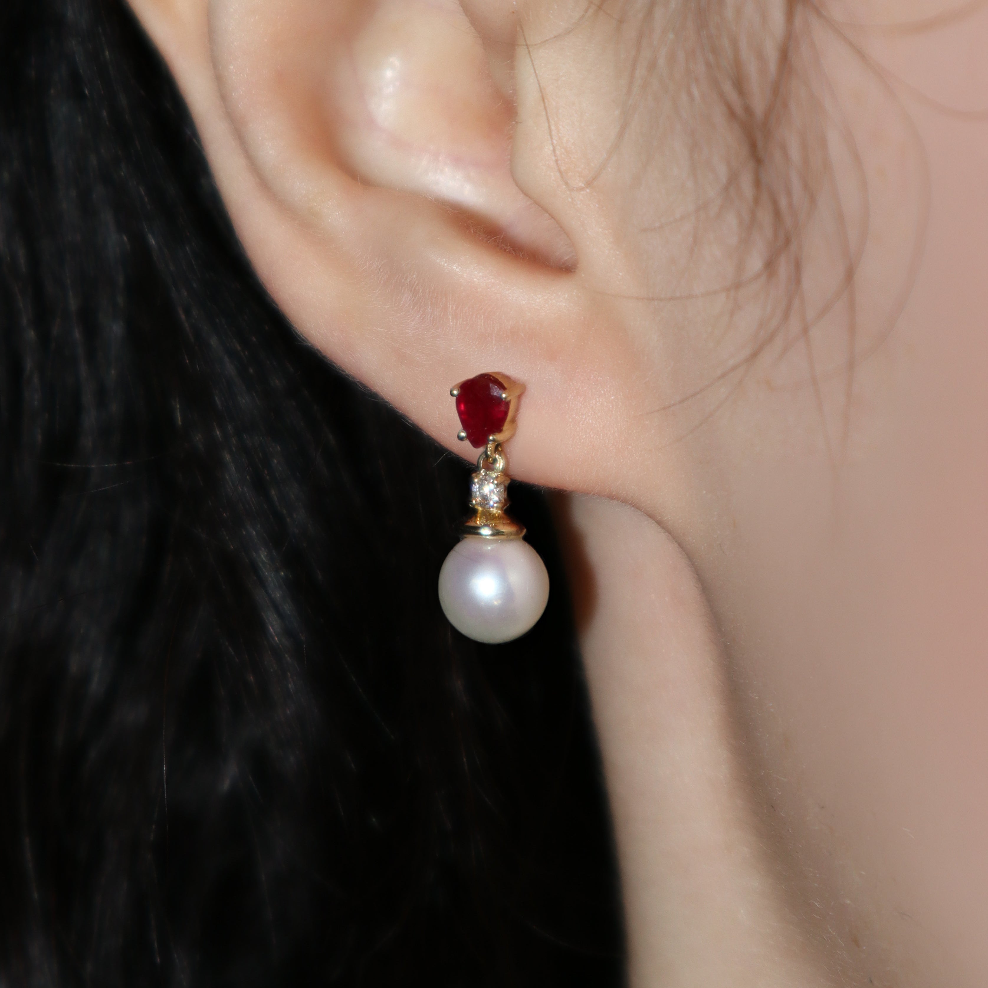 A product photo of a pair of ornate pearl drop stud earrings in yellow gold photographed on a model's earlobe. The pearls are creamy white and round, with one small diamond followed by a pear-shaped ruby on each piece linking them to the back of the golden stud.