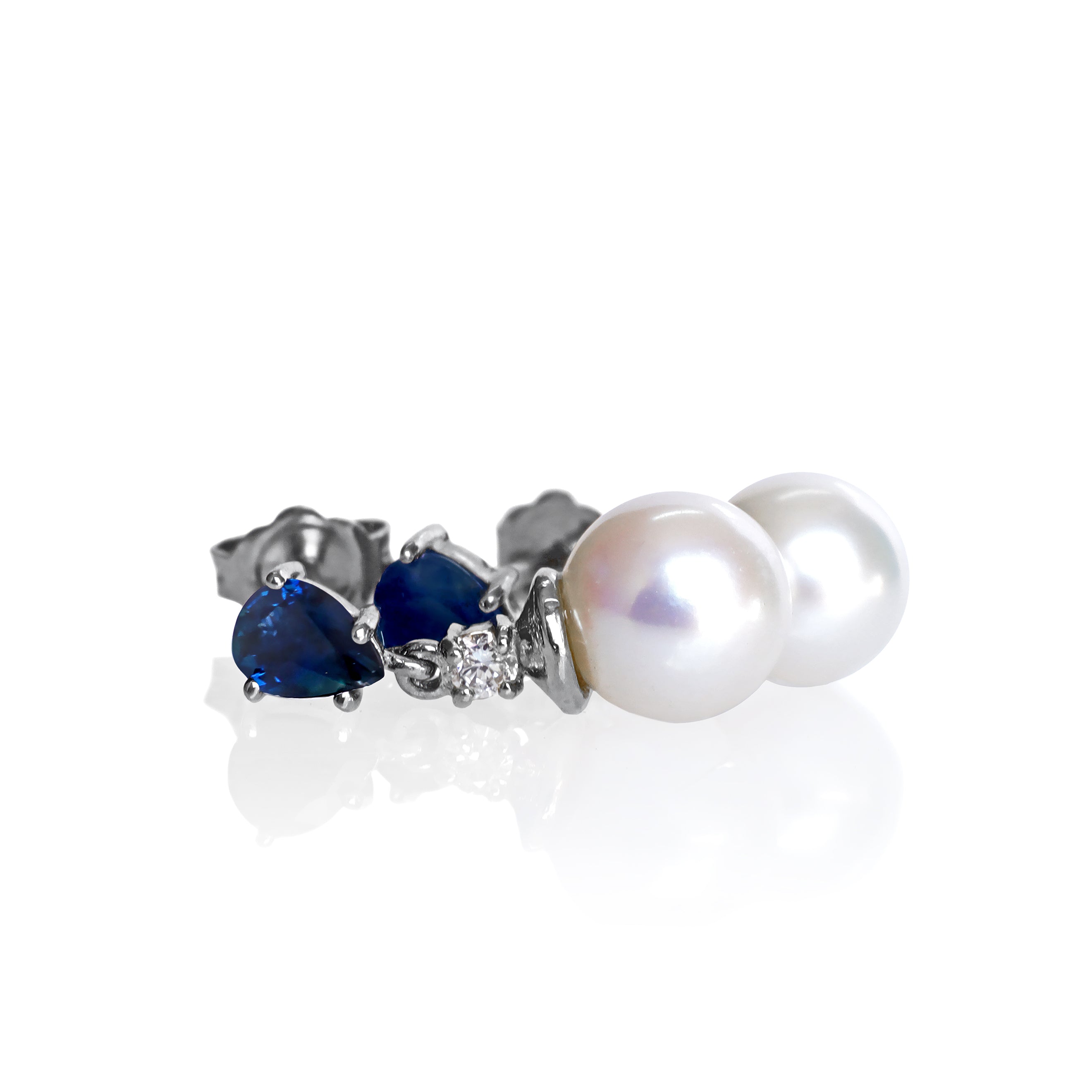 A product photo of a pair of ornate pearl drop stud earrings in white gold on a plain white background. The pearls are creamy white and round, with one small diamond followed by a pear-shaped sapphire on each piece linking them to the back of the golden stud.
