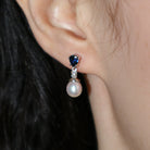 A product photo of a pair of ornate pearl drop stud earrings in white gold photographed on a model's earlobe. The pearls are creamy white and round, with one small diamond followed by a pear-shaped sapphire on each piece linking them to the back of the golden stud.