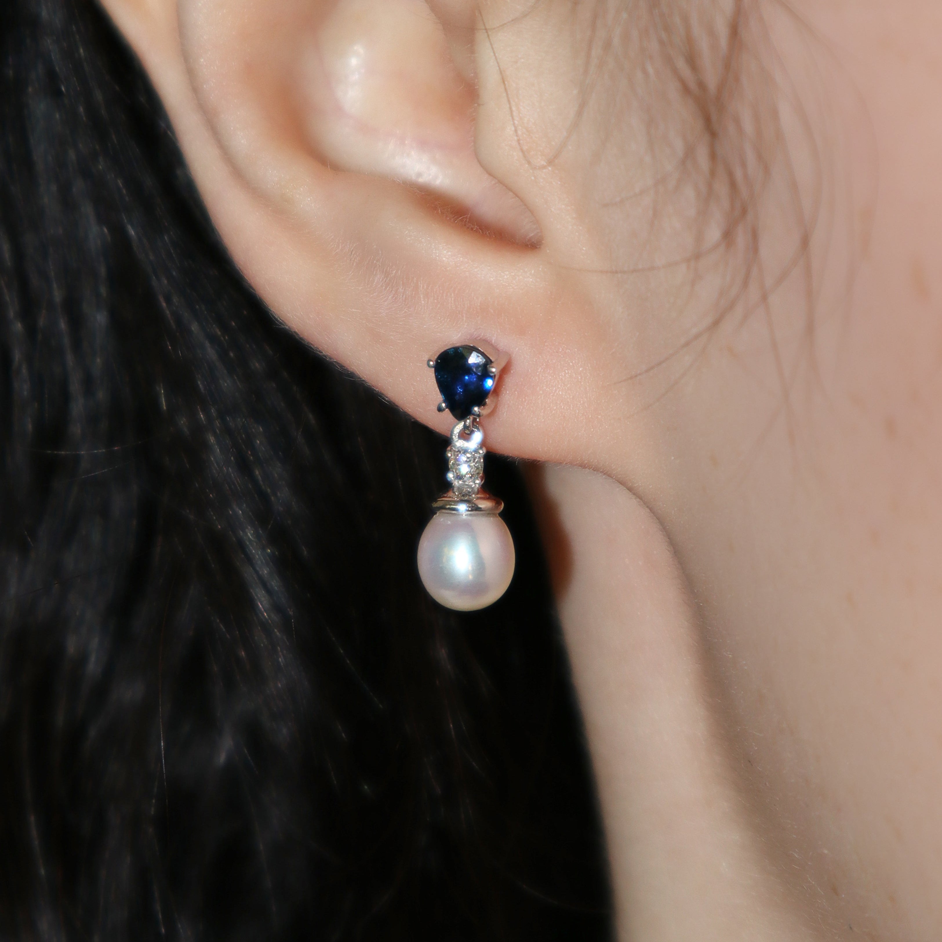 A product photo of a pair of ornate pearl drop stud earrings in white gold photographed on a model's earlobe. The pearls are creamy white and round, with one small diamond followed by a pear-shaped sapphire on each piece linking them to the back of the golden stud.