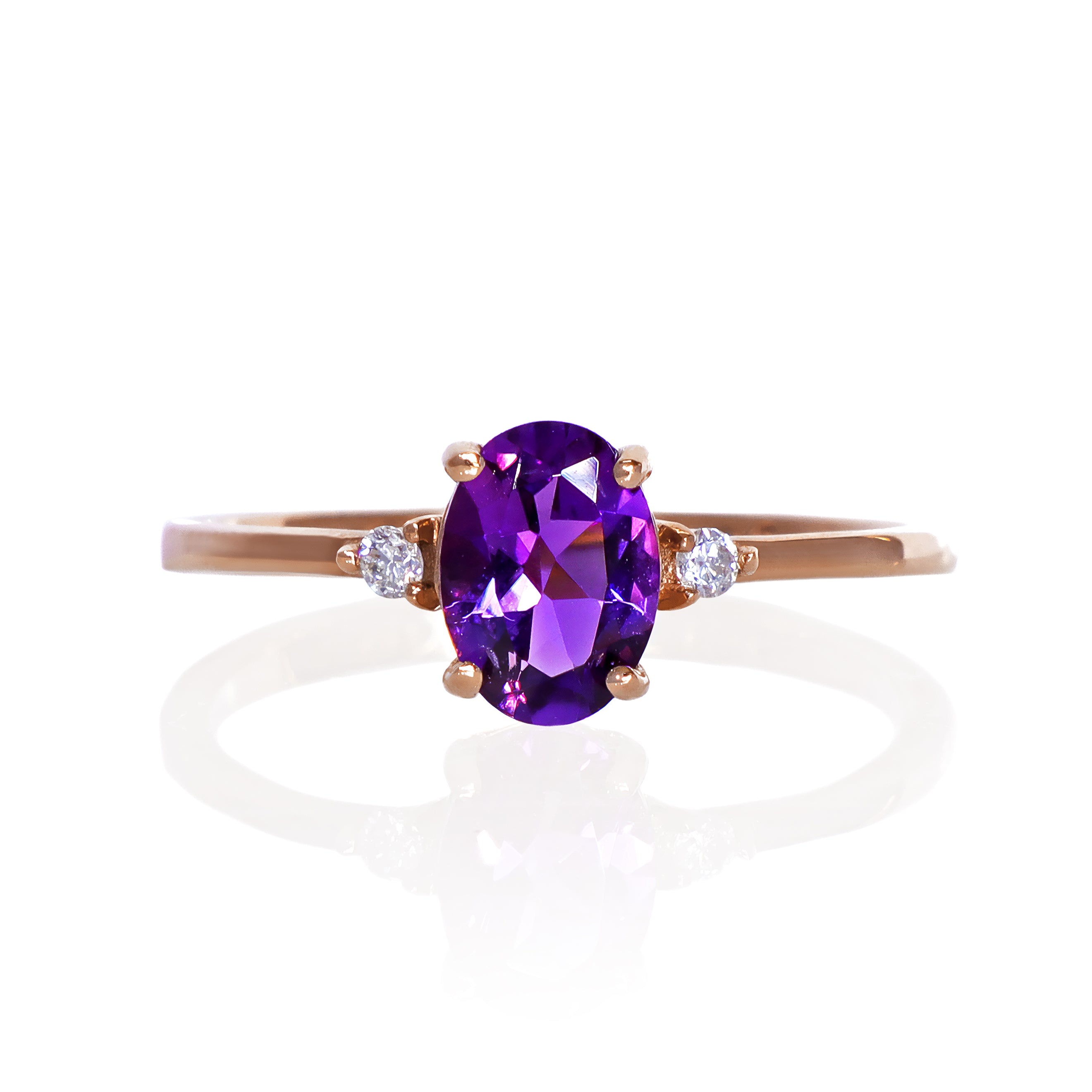 A product photo of a rose gold amethyst ring sitting against a white background. The rose gold band is plain and smooth, and the centre oval-cut deep purple amethyst stone is framed by a single white diamond on either side.