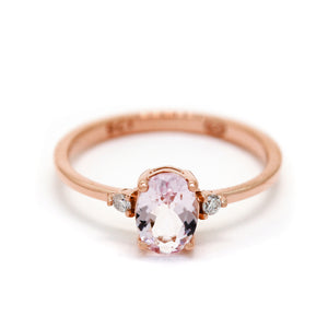 A product photo of a rose gold morganite ring sitting against a white background. The rose gold band is plain and smooth, and the centre oval-cut light pink morganite stone is framed by a single white diamond on either side.