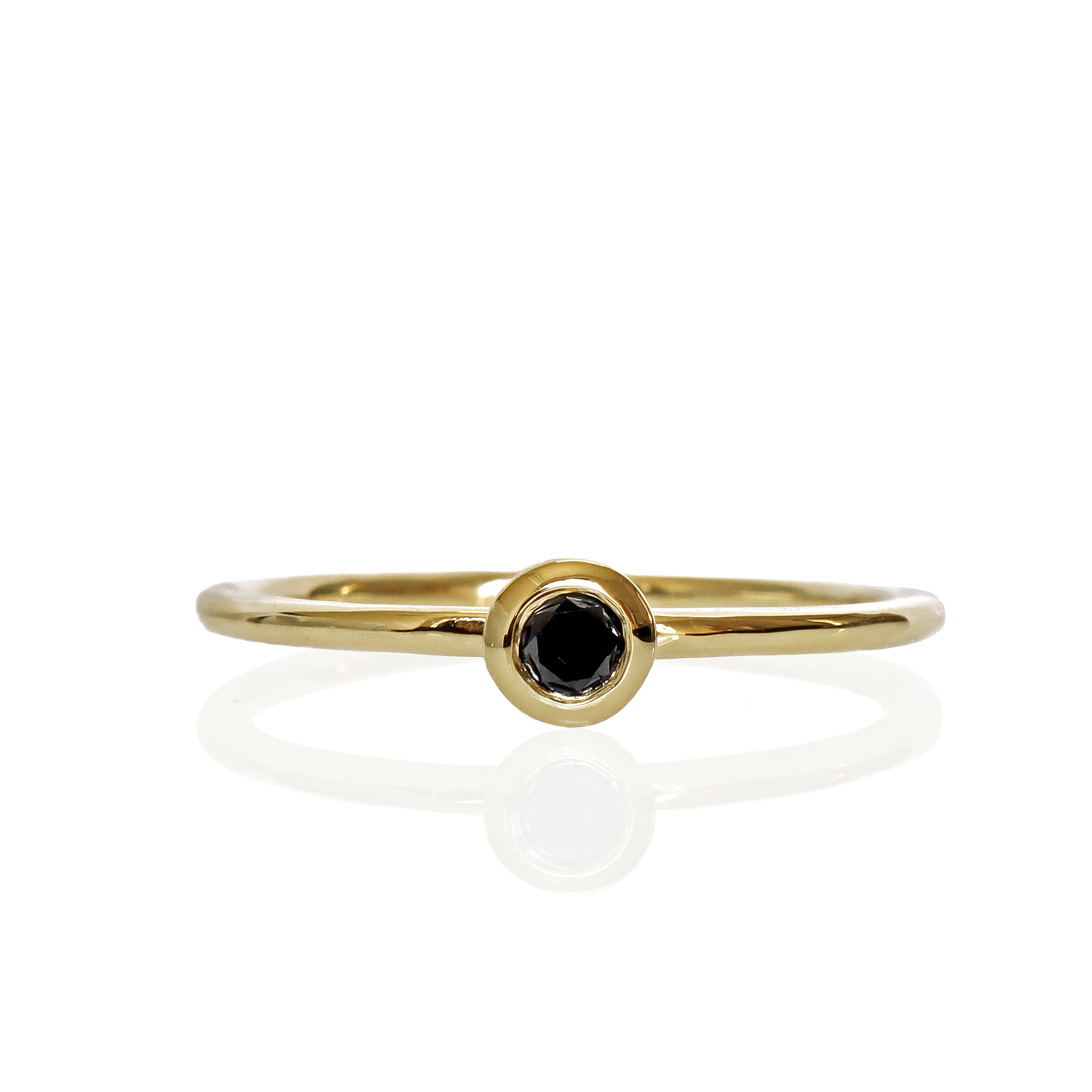 A product photo of a delicate yellow gold stacking ring with a tiny, bezel-set black diamond in the centre sitting on a white background. The band is slim and thread-like, with the focus drawn to the petite 3mm glinting black centre stone.