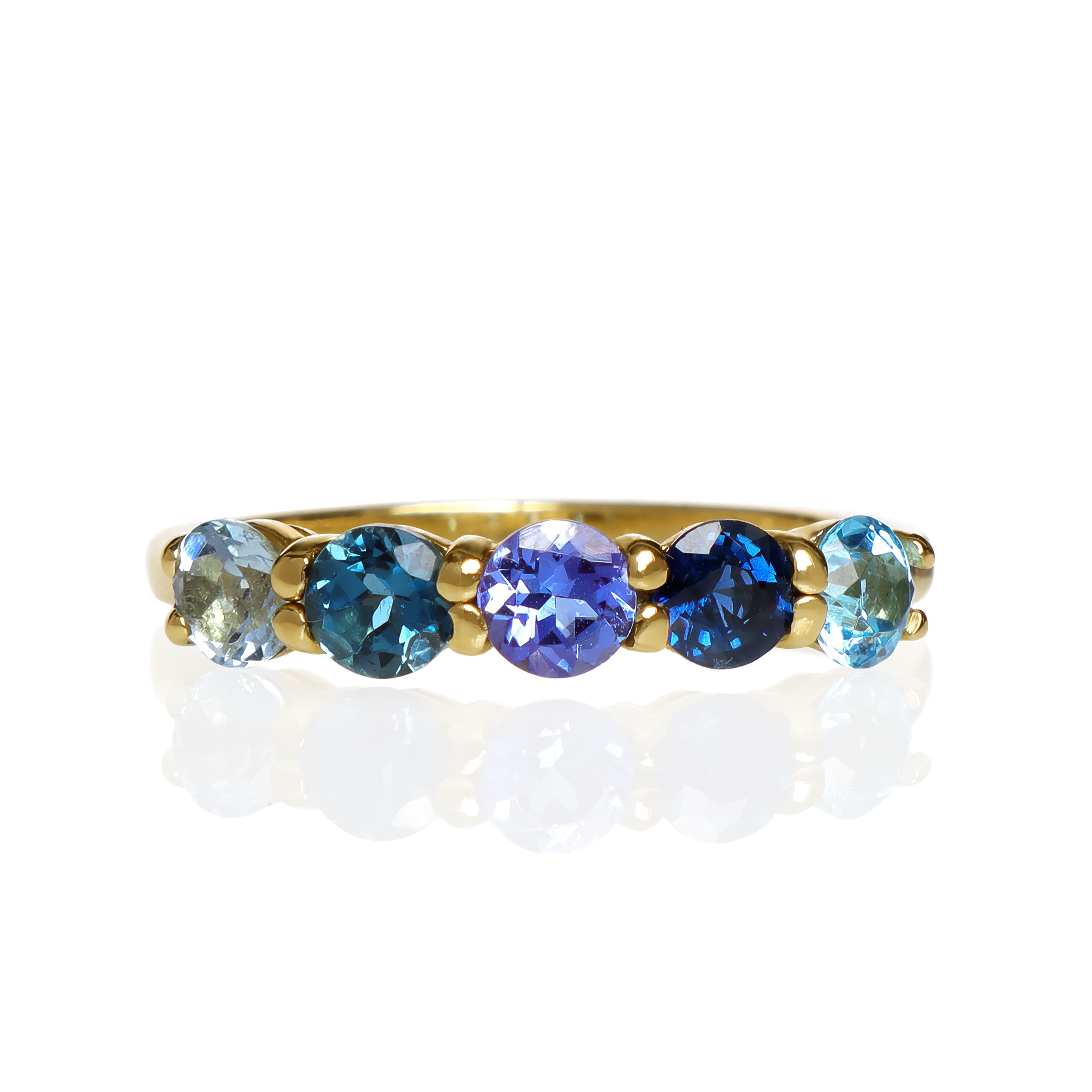 A product photo of a bold, multi-gemstone ring in 9k yellow gold – made up of 5 blue coloured jewels – sitting on a white background. From left to right, 4.5mm round-cut aquamarine, blue tourmaline, tanzanite, sapphire and blue topaz stones make up a bold statement piece.