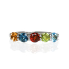 A product photo of a bold, multi-gemstone ring in 9k yellow gold – made up of rainbow-coloured jewels – sitting on a white background. From left to right, 4.5mm round-cut citrine, london blue topaz, red garnet, peridot and blue topaz.