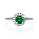 A product photo of a classic diamond halo tsavorite ring in solid 9k white gold on a white background. The diamond halo is framed by a thick band of solid 9k white gold, and the rounded band is smooth and minimalistic. The deep, verdant-green tsavorite centre stone is held in place by 4 claws and reflects light off of its many facets. The green gemstone colour would be a good emerald substitute.