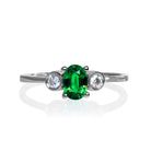 A product photo of a uniquely elegant tsavorite and duo diamond ring in solid 9k white gold on a white background. The deep, verdant-green tsavorite centre stone is held in place by 4 claws and reflects light off of its many facets, while the bezel-set diamonds are framed by a solid layer of white gold. The green gemstone colour would be a good emerald substitute.
