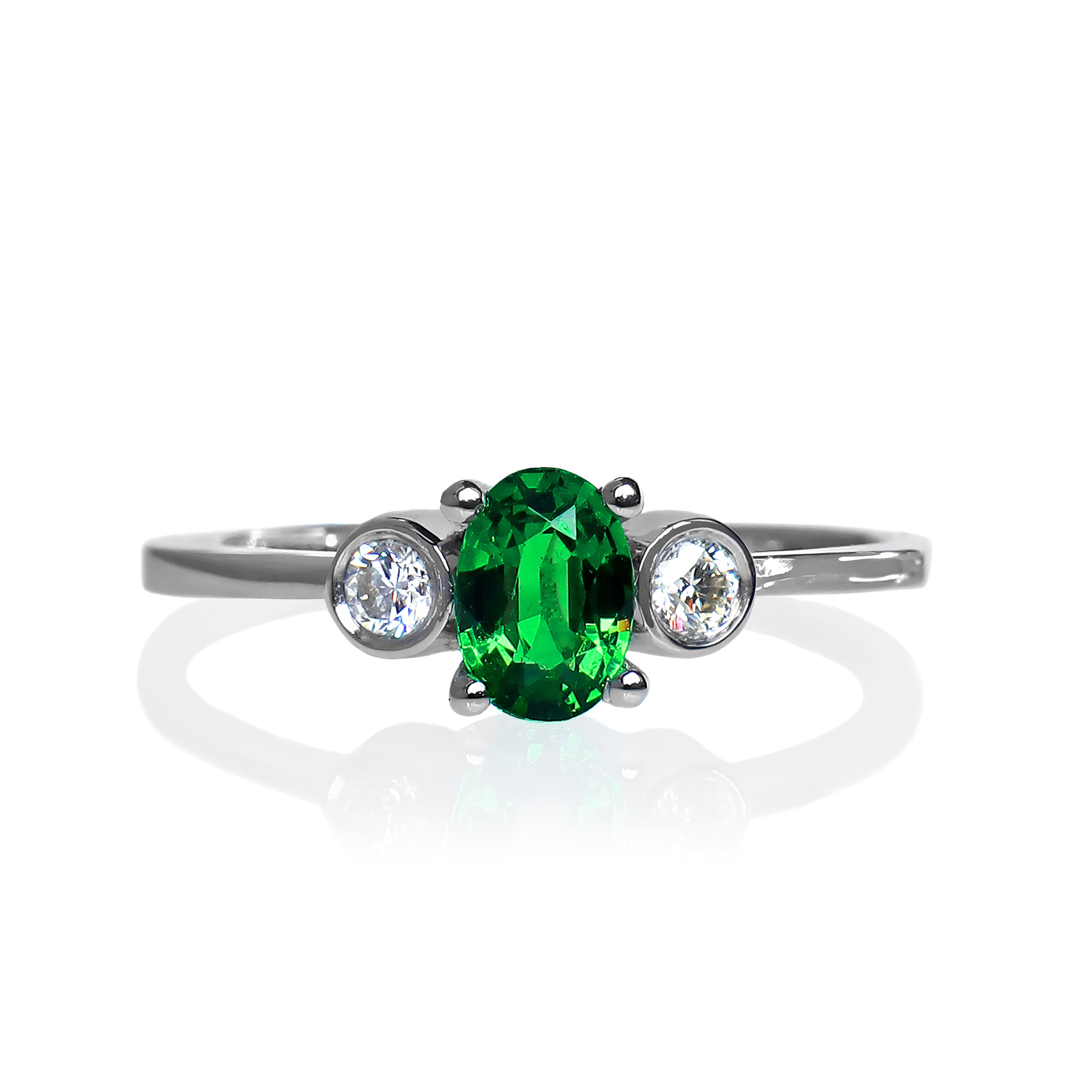 A product photo of a uniquely elegant tsavorite and duo diamond ring in solid 9k white gold on a white background. The deep, verdant-green tsavorite centre stone is held in place by 4 claws and reflects light off of its many facets, while the bezel-set diamonds are framed by a solid layer of white gold. The green gemstone colour would be a good emerald substitute.