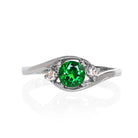 A product photo of a mystical tsavorite and diamond fantasy ring in solid 9k white gold on a white background. The deep, verdant-green tsavorite is held in place by 4 claws and reflects light off of its many facets, while the diamonds on either side are held by 3 each. The smooth white gold band curves elegantly, meeting at the top and bottom of the tsavorite and diamond arrangement. The green gemstone colour be a good emerald substitute.