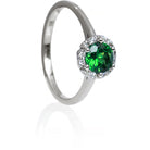 A product photo of a classic diamond halo tsavorite ring in solid 9k white gold on a white background. The rounded band is smooth and minimalistic. The deep, verdant-green tsavorite centre stone is held in place by 6 claws and reflects light off of its many facets. The green gemstone colour would be a good emerald substitute.