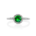 A product photo of a classic diamond halo tsavorite ring in solid 9k white gold on a white background. The rounded band is smooth and minimalistic. The deep, verdant-green tsavorite centre stone is held in place by 6 claws and reflects light off of its many facets. The green gemstone colour be a good emerald substitute.