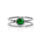A product photo of a split-band white gold ring with a bezel-set tsavorite centre stone sitting on a white background. The band splits at the base of the ring, separating to meet at the top and bottom of the verdant-green tsavorite centre stone, framed in a thick layer of gold, holding it in place.  The green gemstone colour would be a good emerald substitute.