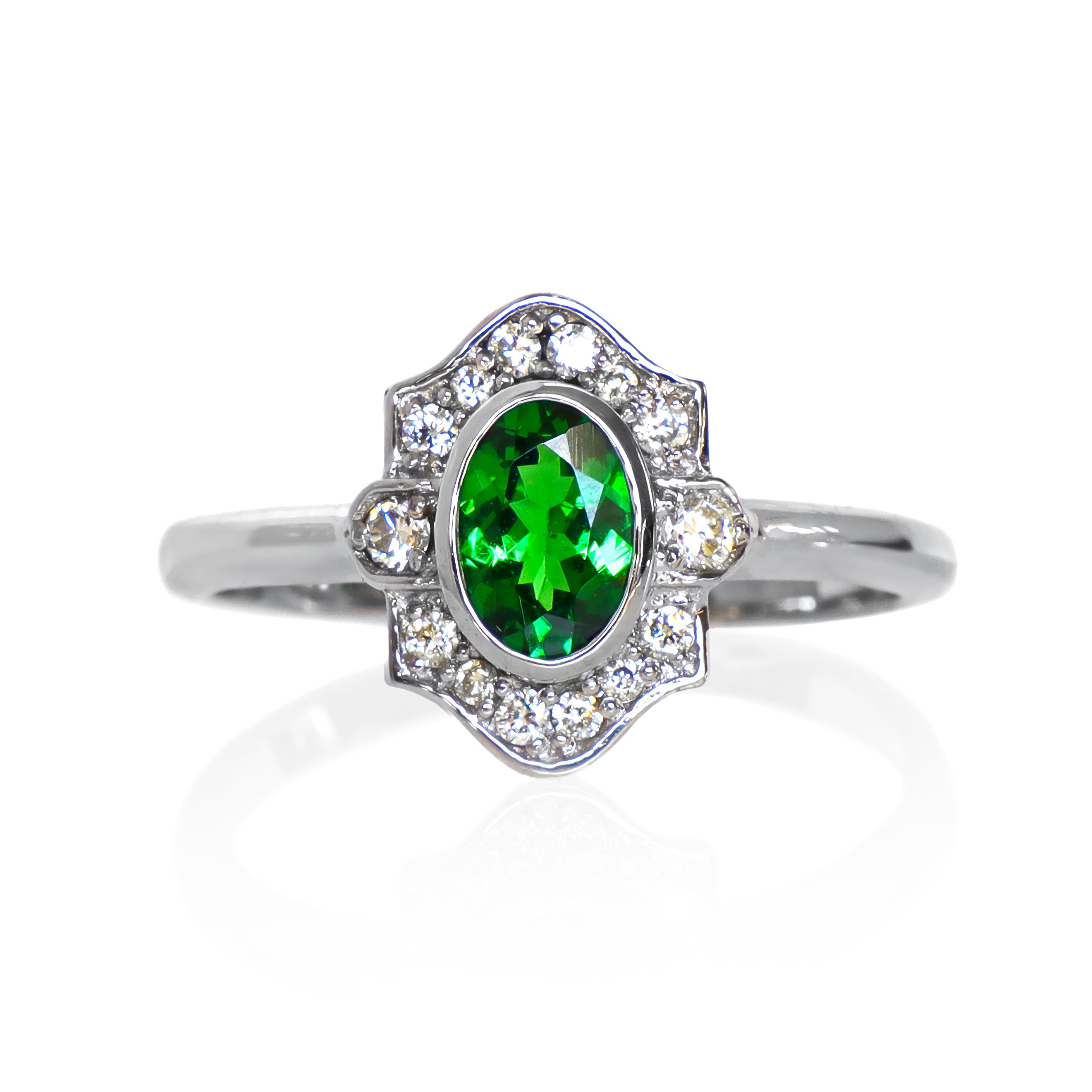 A product photo of a vintage-styled tsavorite and diamond ring in 9k white gold on a white background. The oval-shaped tsavorite is held in place by a bezel setting, and is surrounded by an ornate white gold frame embedded with diamonds, reminiscent of vintage jewellery styles of the past. The green gemstone colour would be a good emerald substitute. 
