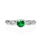 A product photo of an ornate white gold ring with a tsavorite centre stone sitting on a white background. The band splits halfway along its length, becoming twisting and serpentine in appearance before meeting on either side of the deep 5mm round tsavorite, which is held in place by 4 silver claws.  The green gemstone colour be a good emerald substitute.