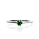A product photo of a delicate white gold stacking ring with a tiny, bezel-set tsavorite in the centre sitting on a white background. The band is slim and thread-like, with the focus drawn to the petite 3mm glinting green centre stone. The green gemstone colour be a good emerald substitute.