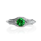 A product photo of an ornate, filigree-detailed white gold ring - reminiscent of an elvish fantasy ring with a deep forest green tsavorite centre stone. The band is detailed with delicate curves and patterns for about half of its length around the dazzling tsavorite stone, before smoothing out towards the back of the ring. The green gemstone colour would be a good emerald substitute.