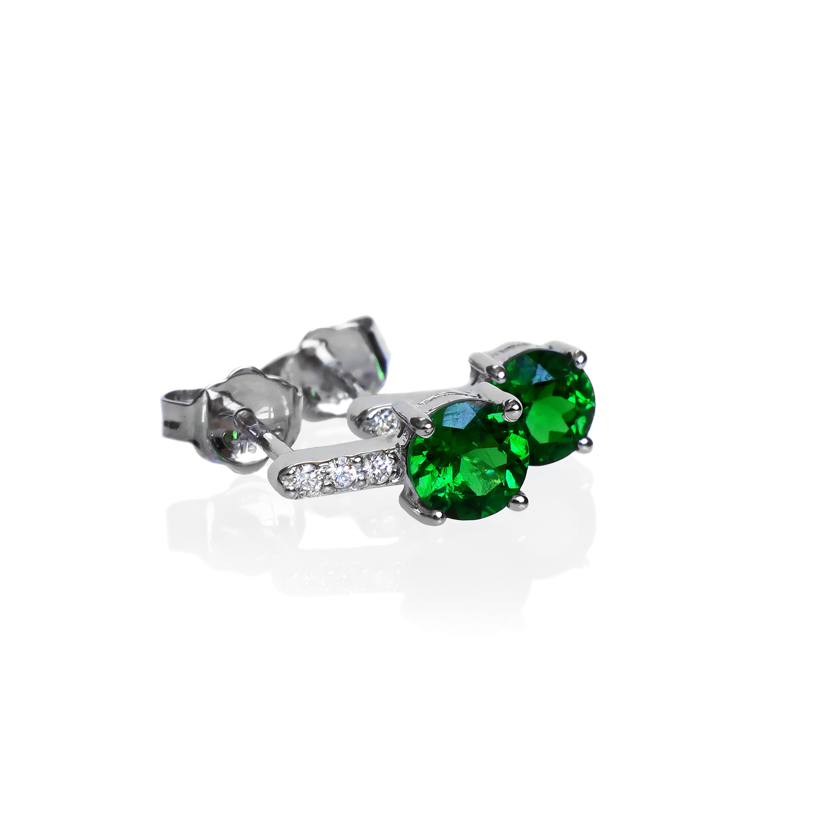 A product photo of a pair of white gold tsavorite earrings sitting against a white background. The simple circle-cut stones are contrasted by the white diamond trio protruding in a straight line above each stone, encased in white gold.